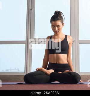 Vista frontale della ragazza brunetta seduta in posa lotos, meditando al chiuso. Donna snella che fa yoga, seduta su gambe incrociate con occhi chiusi sul tappetino yoga. Concetto di equilibrio interiore. Foto Stock