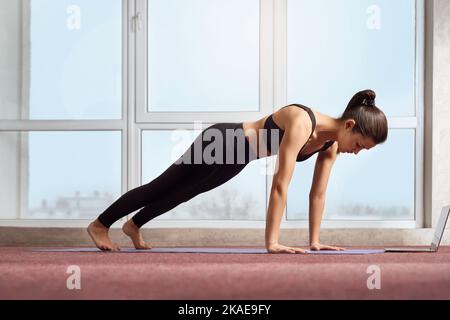 Vista laterale di una giovane donna forte che si esercita con un computer portatile. Donna che indossa tuta sportiva nera, in piedi in tavola, pratica yoga in interni. Concetto di stile di vita sano. Foto Stock