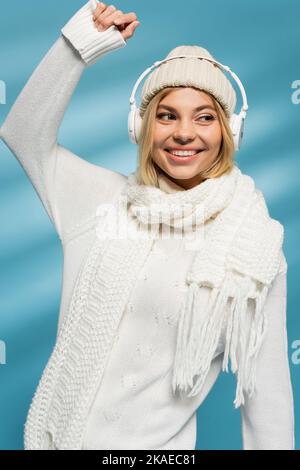 donna bionda felice in cappello invernale e cuffie wireless ascoltare musica su blu, immagine stock Foto Stock