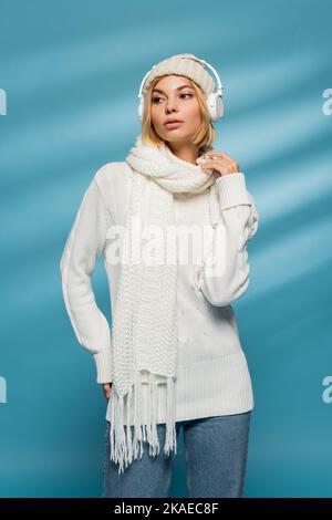 giovane donna bionda in cappello lavorato a maglia e cuffie wireless in posa blu, immagine stock Foto Stock