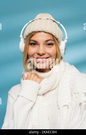 felice giovane donna in maglia cappello e cuffie wireless guardando la fotocamera su blu, immagine stock Foto Stock