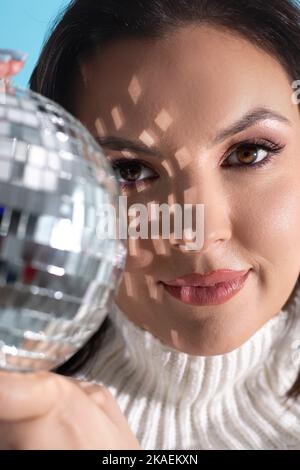 Splendida donna adulta con perfetto trucco tenere in mano splendente palla da discoteca. Primo piano ritratto femminile, riflesso della brillantezza sferica sul volto. Musica retrò Foto Stock