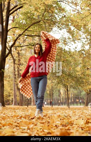 Foto a tutta lunghezza di felice donna allegra in confortevole maglione rosso lavorato a maglia divertirsi con la plaid a scacchi, fogliame dorato sul terreno. Giovane donna entrare Foto Stock