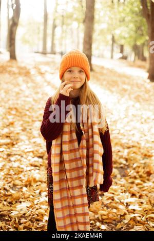 Ritratto di scuola sorridente anziana ragazza isolata su sfondo soleggiato foresta gialla. Bambino felice in caldo cappello, sciarpa e maglione passeggiata nel parco dorato Foto Stock