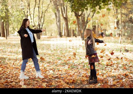 Giovane donna gioiosa e bambina buttare su manciata di foglie gialle asciutte. Felice madre e figlia in abiti neri casual divertirsi e giocare Foto Stock