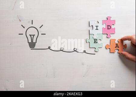 Mano maschio che monta quattro pezzi di puzzle di corrispondenza colorati per incantare la parola idea, collegato a una lampadina disegnata a mano su semplice backgr di legno Foto Stock