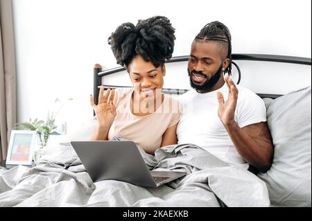 Comunicazione video a distanza. Positivo sposi afro-americani felici, marito e moglie, sono seduti in un letto in camera da letto, utilizzando un computer portatile, parlando in videoconferenza con amici o familiari, sorridendo Foto Stock