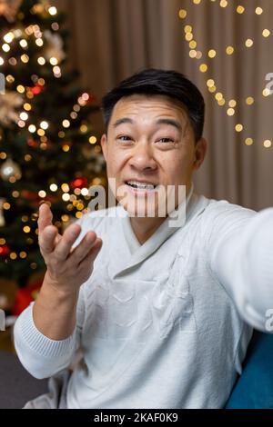 Giovane asiatico che parla sulla fotocamera del telefono come selfie. Si siede a casa vicino all'albero di Natale, saluta, comunica, sorride. Foto Stock