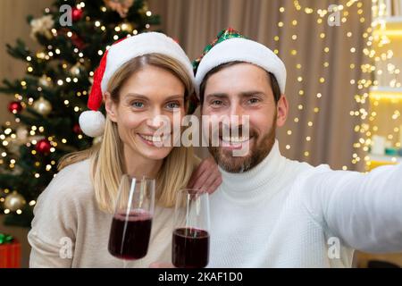 Ritratto festivo di una giovane coppia felice. Sono seduti a casa vicino all'albero di Natale, vestiti con cappelli rossi di Babbo Natale. Stanno tenendo bicchieri di vino, guardando la macchina fotografica, sorridendo Foto Stock