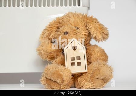 Una lepre di peluche e una piccola casa di legno si trovano vicino al convettore su uno sfondo bianco, una casa calda e un riscaldatore, elettrodomestici di riscaldamento casa Foto Stock