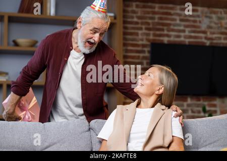 Premuroso vecchio marito dando romantico regalo di compleanno a moglie matura eccitato, amore uomo più anziano sorpresa felice donna di mezza età, scatola rosa aperta, bacio Foto Stock