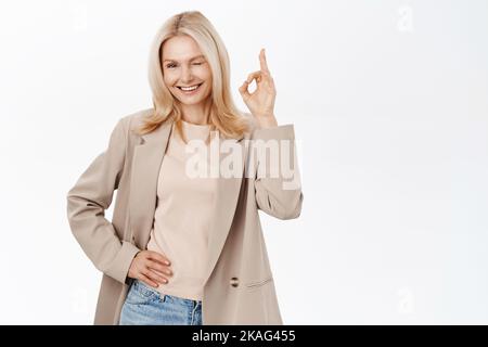Elegante donna anziana, bionda modello femminile nel suo 50s, sorridente e mostrando OK, segno ok, vi lancano, si erge su sfondo bianco. Foto Stock