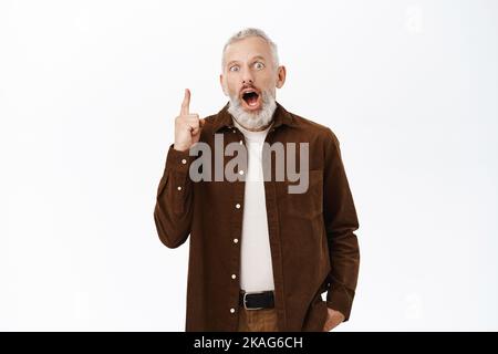 Immagine di un uomo anziano che guarda stupito, puntando il dito verso l'alto a smth emozionante, in piedi su sfondo bianco Foto Stock