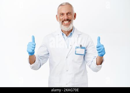 Medico sorridente, medico che mostra i pollici in su, consigliando la clinica, levandosi in piedi sopra lo sfondo bianco Foto Stock