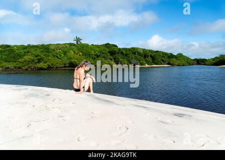 Una donna seduta sul fiume contro la foresta sullo sfondo. Guaibim spiaggia nella città di Valenca, Bahia, Brasile. Foto Stock