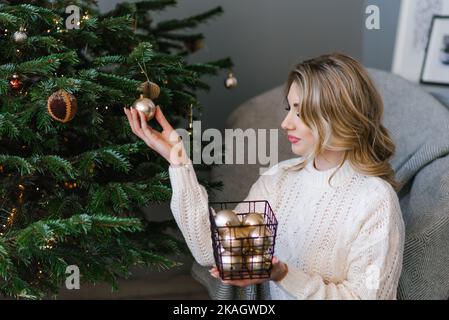 Bella giovane donna in un maglione accogliente e jeans decorare un albero di Natale con palloncini da un cesto a casa Foto Stock