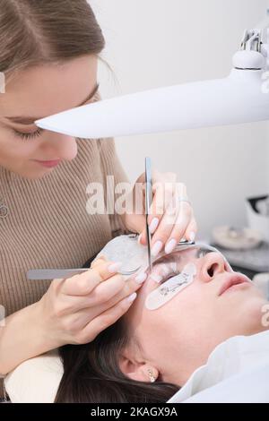 bella donna beautician tenere pinzette con falsa ciglia per incollarlo alla palpebra di un cliente. colpo verticale. processo di correzione del ciglia Foto Stock