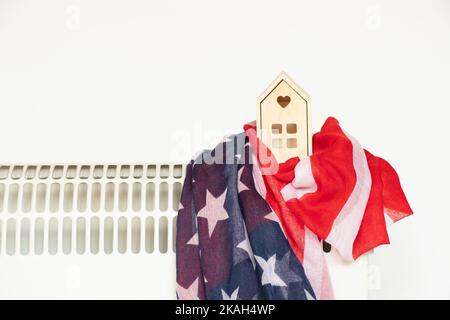 Una piccola casa di legno e la bandiera dell'America si trova su un convettore di riscaldamento su uno sfondo bianco, una casa calda in America, la stagione di riscaldamento Foto Stock