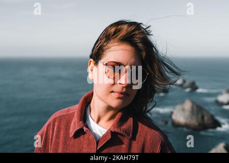 giovane donna caucasica con i capelli da un lato indossando occhiali da sole t-shirt bianca e giacca in corduroy rosso vicino all'oceano in una giornata di sole, punto pepita, nuovo Foto Stock