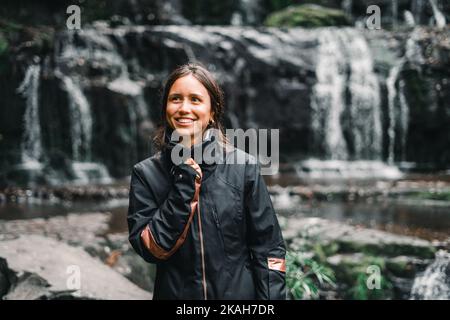 ragazza caucasica con gli occhi neri e la bocca bianca sorridente felicemente e serenamente afferrando la cerniera della sua giacca nera dalle cascate e dal lago nel Foto Stock