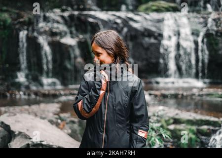 ragazza caucasica con occhi neri e capelli castani felice e calma tenendo la cerniera della sua giacca nera vicino alle cascate e al lago in mezzo alla natura Foto Stock