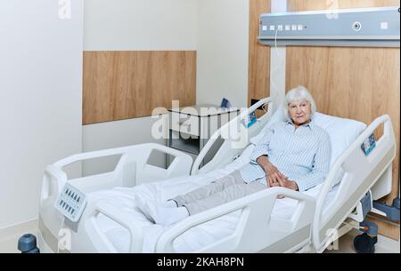 Donna anziana dai capelli grigi sdraiata a letto medico in reparto di un ospedale moderno durante il trattamento. Cura e trattamento delle persone anziane Foto Stock
