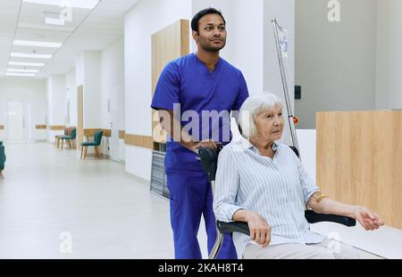 Infermiera maschile indiana che spinge la sedia a rotelle con la donna anziana in corridoio ospedale. Assistenza agli anziani Foto Stock