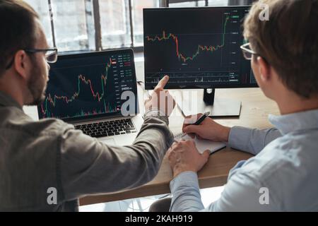 Closeup of two men crypto traders sitting at office table together in front of pc, monitoring stocks data charts on screen, analyzing price flow. Profit teamwork. Digital money Foto Stock