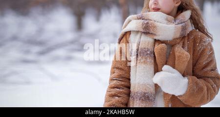 Una ragazza bionda bianca in un cappello a maglia, una sciarpa lunga e un cappotto di pelliccia cammina attraverso la foresta e sorride. Donna cattura la neve in muffole bianche. L'inverno è sulla strada. Foto di alta qualità. Foto Stock