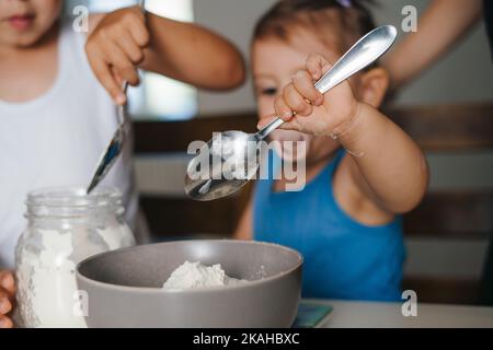 Vista ravvicinata delle mani dei bambini che tengono un cucchiaio e aggiungono la farina al recipiente con gli ingredienti. Nutrizione. Stile di vita sano. Cibo equilibrato. Famiglia Foto Stock