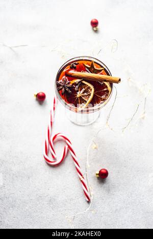 Bicchiere di VIN brulé con fette di arancia secca, bastoncini di cannella e anice stellato accanto a luci a corda, canne caramellate e baubles natalizi Foto Stock