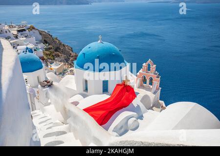 Bellissima ragazza elegante con un lungo vestito rosso che si posa sul tetto bianco della chiesa di Santorini. Felice a Oia, Santorini, isola della Grecia. Tour delle ragazze spensierato Foto Stock
