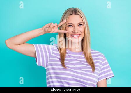 Foto di buon umore ottimista bella donna con capelli dritti abbigliamento a righe T-shirt con segno V isolato su sfondo di colore blu Foto Stock