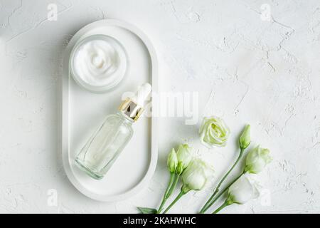 Set di cosmetici naturali anti-età per la cura della pelle su vassoio bianco con fiori di eustoma. Olio viso di bellezza, olio essenziale e crema viso in un bicchiere senza marchio pac Foto Stock