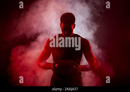 Lato giocatore di basket illuminato con colore rosso che tiene una palla contro sfondo fumo. Uomo africano americano concentrato serio. Foto Stock