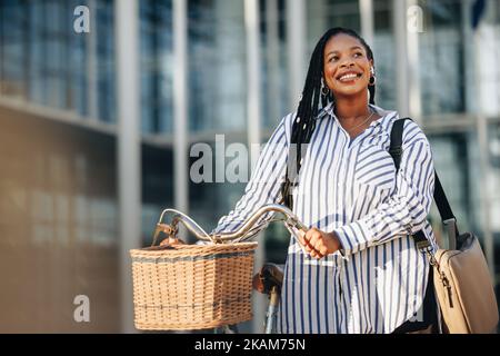 Donna sorridente d'affari che guarda via pensivamente mentre cammina accanto alla sua bicicletta in città. Allegra giovane donna d'affari che si collega al suo ufficio i Foto Stock