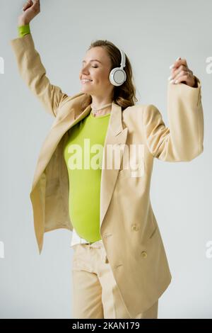 Allegra donna incinta in cuffie e giacca in piedi isolato su grigio, immagine stock Foto Stock