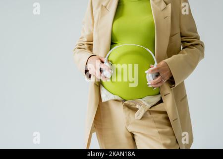 Vista ritagliata di una donna incinta in tuta con cuffie isolate in grigio, immagine stock Foto Stock