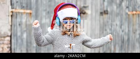 Banner orizzontale o testata con divertente bambina che indossa cappello Babbo Natale e occhiali da sole che ballano su uno sfondo di legno all'aperto in Natale ti Foto Stock