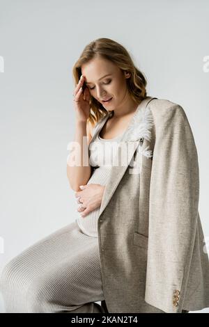 Giovane donna incinta in vestito e giacca guardando la pancia isolato su grigio, immagine stock Foto Stock