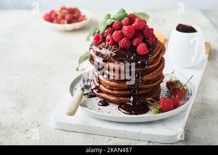 Pancake al cioccolato. Frittelle con lampone fresco con glassa al cioccolato o condimenti in recipiente grigio su sfondo grigio chiaro tavolo. Ameri classici fatti in casa Foto Stock