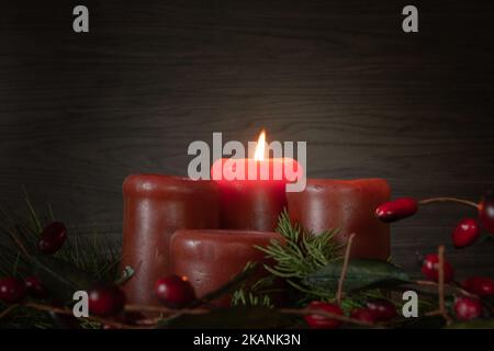 Una candela rossa che brucia in una corona dell'avvento con tosse sempreverde e bacche rosse su uno sfondo scuro con spazio di copia Foto Stock