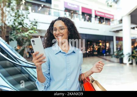 Donna happy hispanic in supermercato shopping, lettura di messaggi online, tenendo borse shopping colorato e smartphone. Foto Stock