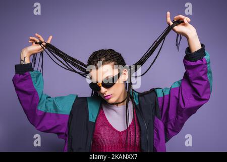 donna alla moda in occhiali da sole e giacca vintage stile che tiene intrecciato dreadlocks isolato su viola, immagine stock Foto Stock