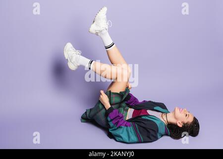 donna in stile anni '90 in sneakers bianco sdraiato con gambe rialzate su sfondo viola, immagine stock Foto Stock