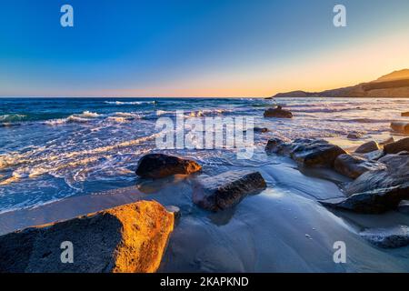 Bella stagione di onde basse maree, cielo limpido tramonto, colline nebulose. Acque azzurre e azzurre che si avvicinano alle pietre lavate con poca schiuma bianca. Foto Stock