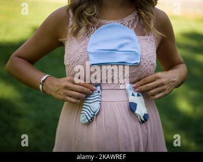 Madre incinta che tiene un cappuccio e due coppie di calzini piccoli del bambino sullo stomaco - concetto di gravidanza. Foto Stock