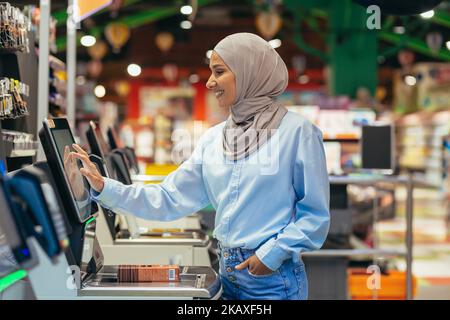 Una donna compratrice in un supermercato in un hijab paga le merci ad un checkout self-service, servizio conveniente per i clienti. Foto Stock