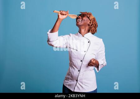 Chef professionista afro-americano che indossa uniforme cucina cantando eccitatamente con cucchiaio di legno. Cuoca femminile che si rilassa dopo il lavoro in studio girato su sfondo blu. Foto Stock