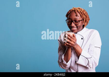 Rilassato cuoca femminile gustando una tazza di caffè preferito durante l'ora di pranzo. Chef professionista afroamericano che indossa uniforme da cucina su sfondo blu in studio shot. Foto Stock